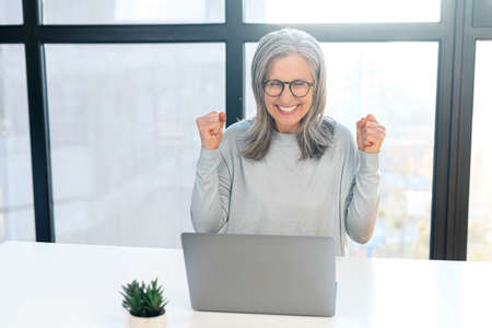Overjoyed mature woman in casual wear with laptop in office, watching at the screen and screaming happily, excited senior lady got job hiring, get good news or won in lottery. Victory conceptの写真素材
