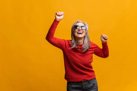 Excited enthusiastic mature woman in sunglasses dancing isolated on yellow, crazy overjoyed senior...
