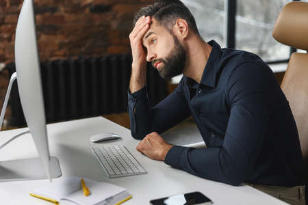Depressed young male office employee is staring at computer monitor with despair, worried businessman made mistake in project, fail deadlineの写真素材