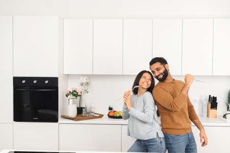 Portrait of joyful married couple laughing at home. Woman and her handsome man smiling and having fun together, while holding kitchen utensilsの写真素材