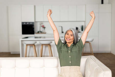 Gray-haired woman stretching arms sitting on the sofa at home, cheerful smiling Asian lady preparing doing stretching exercise, taking a breakの写真素材