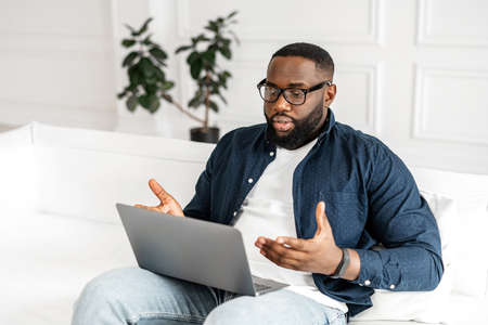Serious concentrated African-American male student involved virtual meeting, webinar, black man making video call on the laptop, sitting on the sofa at home, talking and gesturing, explaining smthの写真素材