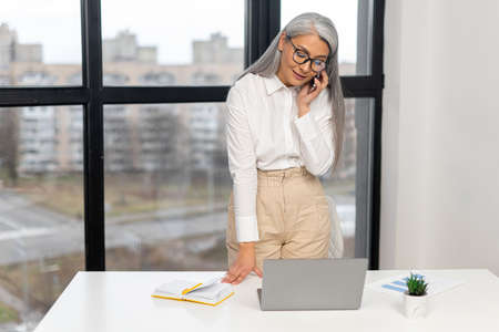 Serious old senior business woman wearing smart casual shirt and stylish eyeglasses standing and chatting via smartphone in modern office space. Mature bossy lady, elderly purposeful leader conceptの写真素材