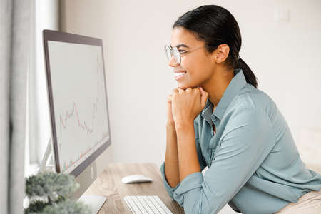 Side view of a biracial girl sitting with computer at the table at cozy living room. Young woman typing on keyboard, messaging with friends onlineの写真素材
