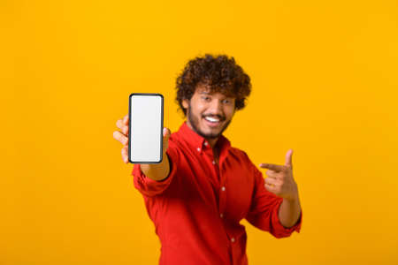 Portrait of satisfied brunette man with curly hair pointing at cellphone and smiling at camera,...
