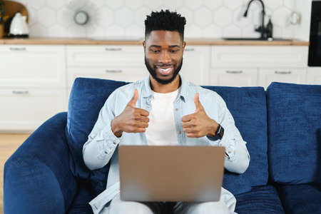 Cheerful African-American guy taking a part in virtual meeting sitting with a laptop on laps on sofa...