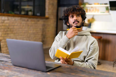 Male student pondering and looking away while writing lecture in a notebook at home. He using laptop...