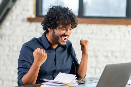 Excited Indian man sitting at the table, looking at the laptop screen and rejoicing at good news....