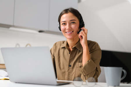 Smiling asian female freelancer wearing headset with microphone using laptop for online connection with colleagues or customers working remotely, woman sitting in the home office ad looks at cameraの写真素材