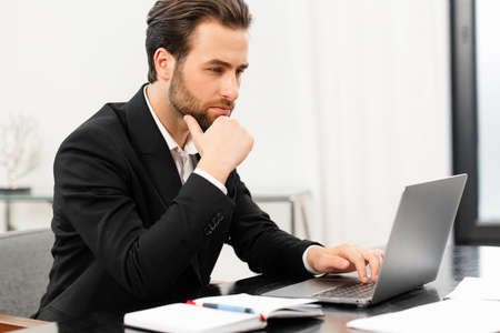 Handsome focused young businessman in formal wear using laptop computer sitting at the desk in...