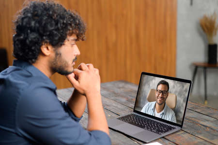 Back view of the Indian guy talking with male friend or coworker via video call. He sitting at the office desk and using laptop for video meeting. Young man on the screenの写真素材