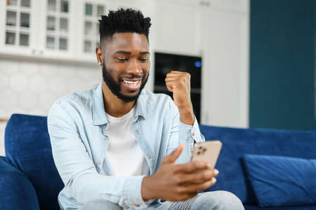 Excited black multiracial man sitting on the sofa looking at the phone and smiling, surprised emotional lucky man, rejoicing at good message, news, won the lotteryの写真素材