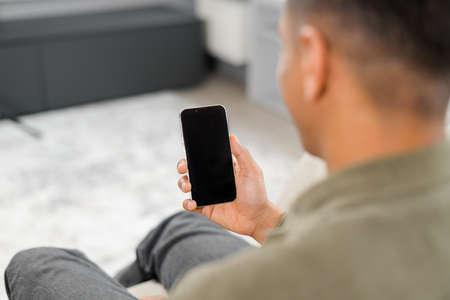 Back view of multiracial man sitting on sofa in office, using mobile phone for checking social nets,...