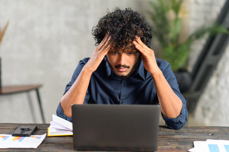Young tired bearded office worker freelancer in casual clothes sitting at the desk at his workplace,...