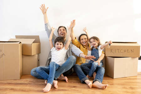 Overjoyed multiracial family of four sitting on floor surrounded cardboard boxes in empty living...