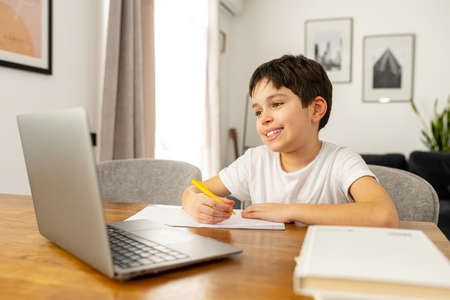Smiling schoolboy studying on the distance from home, using trendy laptop, preteen boy doing...