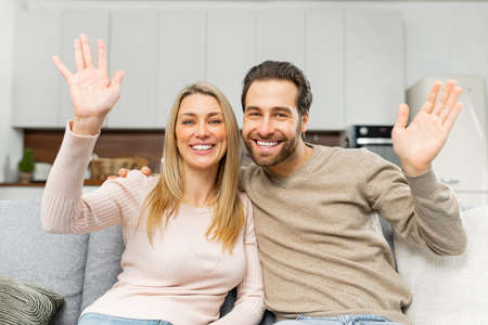 Video screen with cheerful caucasian couple looking at the camera and waving. Webcam view of smiling woman and her man sitting on the couch and greeting. Virtual meeting conceptの写真素材