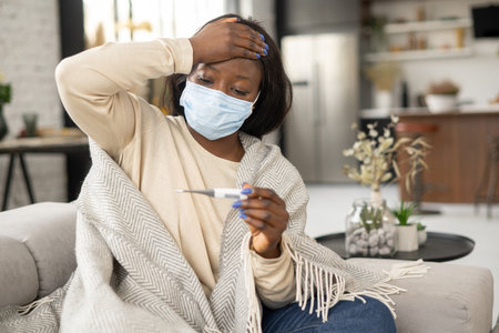 Sick upset african-american woman wearing mask measuring temperature sitting at home. Ill black lady with cold or flu fells fever and suffering from viral disease, self isolation conceptの写真素材