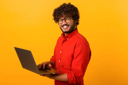 Portrait of young multiracial guy standing isolated on orange wall and holding laptop, typing...