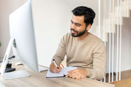Concentrated positive Indian man thinking, taking notes while sitting at the table with computer at...