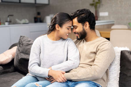 Happy Indian newlyweds moved in new apartment, sitting at the sofa in embraces, looking at each...