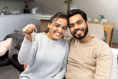 Young married couple smiling cheerfully and showing keys from a new apartment, hugging and looking at camera, standing in the modern kitchen of a new home, real estate and family conceptの写真素材