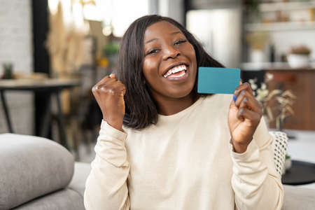 Happy young woman smiling with delight, while sitting at the sofa with credit card and looking at the camera. Happy biracial girl showing plastic credit card. Online shopping conceptの写真素材