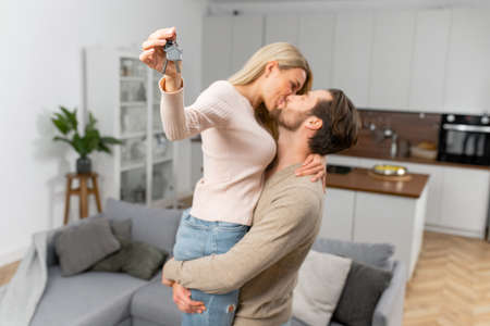 Woman holding home keys while kissing with her boyfriend in new apartment. Lovely couple feeling ecstatic about their recent purchase of new home which makes them feel being on the verge of happinessの写真素材