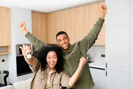 Young multiracial married couple smiling cheerfully and showing keys from a new apartment, hugging and looking at camera, while standing in modern kitchen of new home. Real estate and family conceptの写真素材