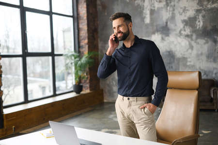 Smiling successful contemporary man manager in casual wear holding phone call. Happy inspired caucasian male small business owner talking on the smartphone while standing in modern loft officeの写真素材