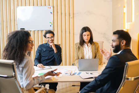 Multiracial team. Group of ambitious young people in smart casual wear discussing business tasks while sitting at the table in a modern office. Flip chart on the backgroundの写真素材