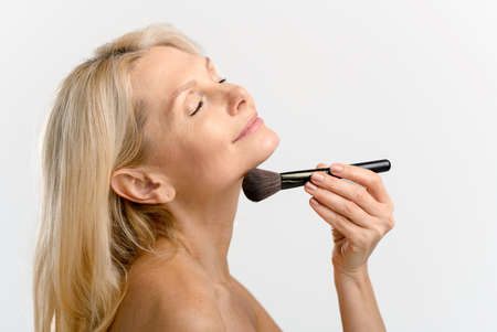 Profile of beautiful 50s middle-aged woman holding makeup brush near chin and applying powder, eyes closed dreamly, charming lady isolated on white background. Skincare conceptの写真素材