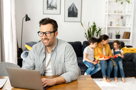 Happy father wearing glasses is working on the distance. Man using laptop for freelance work sitting...