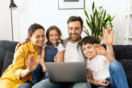 Virtual meeting. Family of four waving to screen while using laptop for video connection with...