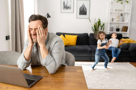 Annoyed father sitting with laptop with eyes closed, covering head with hands while his two kids are jumping and shouting around. Irritated freelancer parent is babysitting and workingの写真素材