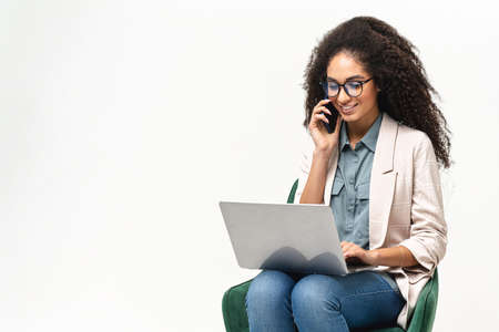 Confident African-American woman in smart casual wear using laptop and talking on the smartphone...