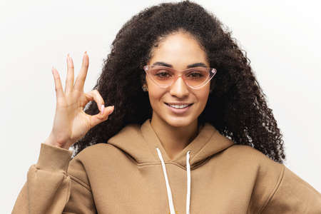 Adorable afro-american woman in casual hoodie and pink sunglasses showing ok sign with fingers...