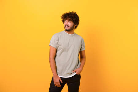 Portrait of happy satisfied curly Indian young man in t-shirt standing and looking away with light...