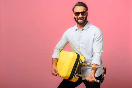 Overjoyed joyful Indian guy in sunglasses stands with a yellow suitcase, plays on a bag like a...