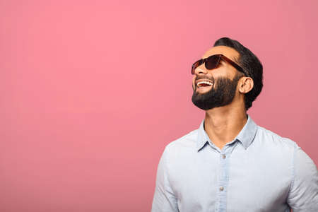 Cheerful Indian man in sunglasses laughing toothy, carefree hispanic guy in blue shirt standing and laughs out loud over pink background. Happy lifestyleの写真素材