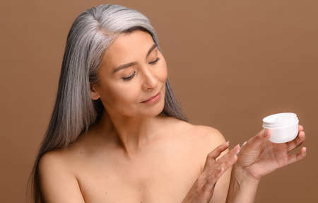 Well-looking middle-aged Asian woman with grey hair testing new cosmetology product, standing and...