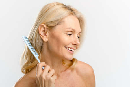 Elegant serene middle-aged blonde woman combing with crest, looks down and laughing cheerfully...