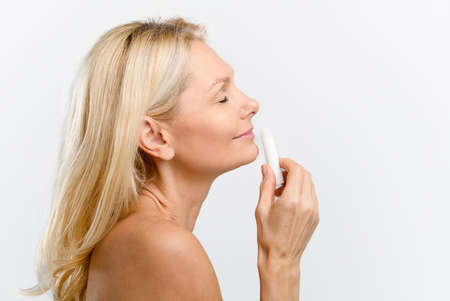 Side view at charming middle-aged woman holding hygienic lipstick, testing and advertising new...