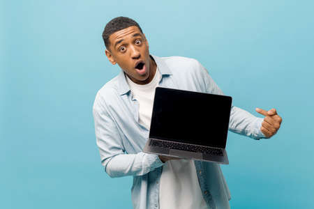 Surprised male student in casual shirt points finger at laptop pc screen with empty display isolated on blue background, shocked multiracial freelancer man dazed by possibilities of new app, mockupの写真素材