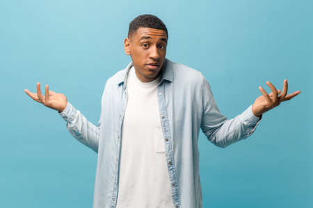 Confused multiracial guy in casual shirt shrugging shoulders isolated on blue background, uncertain...