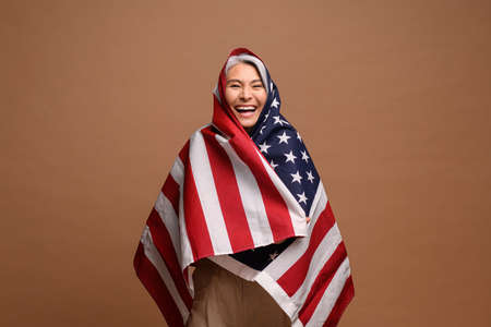 Portrait of mature woman wrapping at the USA flag isolated on brown background. Overjoyed senior...