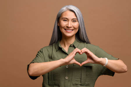 Love you. Portrait of attractive romantic mature woman standing and making heart with hands, while smiling playfully. Indoor studio shot isolated on brown backgroundの写真素材