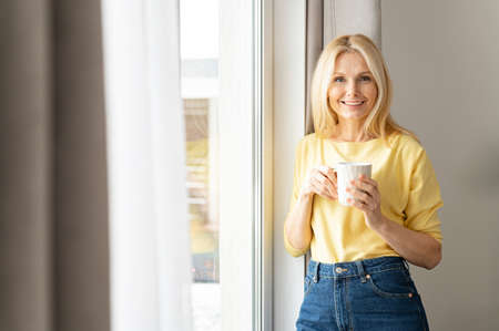 Charming elderly woman stands with cup of morning coffee looks at the camera and smiles, friendly...