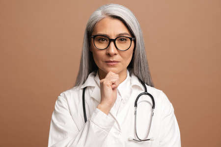 Serious middle-aged Asian female doctor with a stethoscope in a medical coat posing isolated over brown background, looking for solution or thinking about something, resting chin on handの写真素材