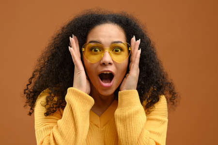Shocked and amazed young attractive woman looks at the camera isolated on brown, a curly multiracial girl screams surprised, holding cheeks, incredible sales or dealの写真素材
